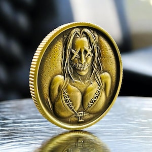 May include: A gold-colored coin featuring a stylized female figure with a skull face and long hair. The figure is wearing a bikini top. The coin has a ridged edge.