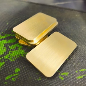 Könnte beinhalten: Mehrere rechteckige, goldfarbene Metallplatten mit abgerundeten Ecken sind auf einer dunklen Oberfläche gestapelt und angeordnet. Die Platten haben eine gebürstete Metallstruktur, und die Beleuchtung hebt ihren metallischen Glanz hervor.