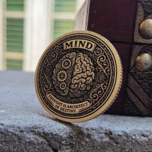 Puede incluir: Una moneda dorada con la palabra "MIND" en la parte superior. La moneda está dividida en dos mitades, una con engranajes y la otra con un diseño de cerebro. El texto "THOUGHT IS ARCHITECT OF DESTINY" está en el borde inferior.