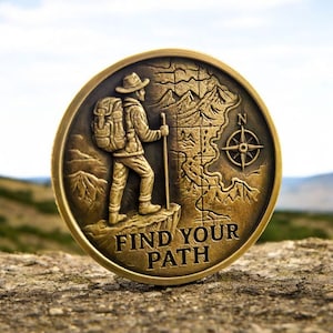 Pode incluir: Uma moeda de cor bronze com um caminhante com uma mochila e um bastão, em pé num penhasco. Um mapa e uma bússola estão no lado direito. As palavras "FIND YOUR PATH" estão na parte inferior.