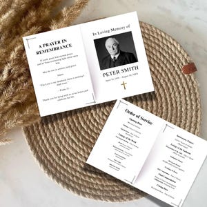 Könnte beinhalten: Zwei weiße Gedenkgottesdienstprogramme mit schwarzem Text. Auf einem Programm steht „Ein Gebet zum Gedenken“ und auf dem anderen „In liebevoller Erinnerung an Peter Smith“. Ein goldenes Kreuz ist sichtbar. Die Programme liegen auf einem gewebten, kreisförmigen Platzset.