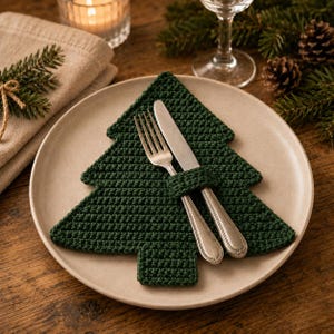 Pode incluir: Um suporte para talheres em crochê verde-escuro em forma de árvore de Natal sobre um prato redondo bege. O suporte possui uma pequena alça integrada que mantém juntos um garfo e uma faca de metal prateado no centro da árvore.