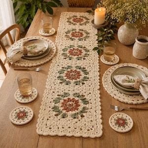 May include: A cream-coloured crochet table runner featuring a repeating floral motif in terracotta and sage green. The set includes matching round crochet placemats and circular coasters, all crafted with a scalloped edge and intricate yarn work.