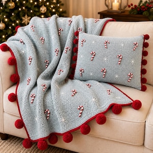 Könnte beinhalten: Eine hellblaue gehäkelte Wohndecke und ein passendes rechteckiges Kissen, beide verziert mit kleinen rot-weißen Zuckerstangenmotiven und weißen Sternakzenten. Die Ränder sind mit einer leuchtend roten Borte und großen roten Bommeln versehen.