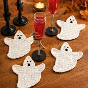 May include: Four white crocheted ghost coasters with black stitched eyes and mouths. The coasters are arranged around two black candle holders, a glass candle, and two tall glasses filled with red liquid, one held by a skeletal hand stem.