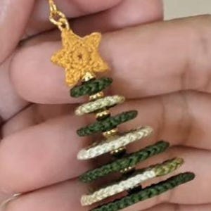 May include: A dangle earring featuring a gold-toned metal hook and a crocheted yellow star at the top. Below the star, the earring is constructed from alternating dark green and cream-colored braided rings, creating a tiered Christmas tree shape.