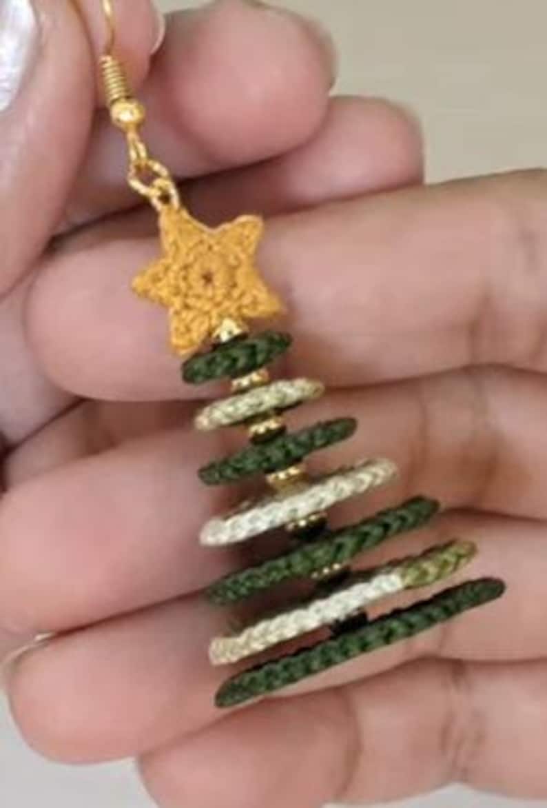 May include: A dangle earring featuring a gold-toned metal hook and a crocheted yellow star at the top. Below the star, the earring is constructed from alternating dark green and cream-colored braided rings, creating a tiered Christmas tree shape.