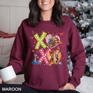 May include: Maroon sweatshirt featuring the word "XO" in a playful design with cartoon characters inside heart shapes. The sweatshirt has a crew neck and long sleeves. The design includes a snake, hearts, and other small graphics.
