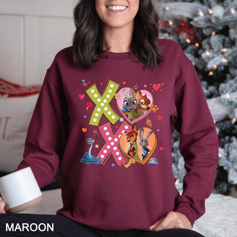 May include: Maroon sweatshirt featuring the word "XO" in a playful design with cartoon characters inside heart shapes. The sweatshirt has a crew neck and long sleeves. The design includes a snake, hearts, and other small graphics.