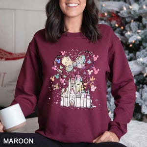 May include: Maroon sweatshirt featuring a colorful graphic of a castle and Minnie Mouse ears. The design includes pastel colors and white speckles. The word "MAROON" is printed at the bottom of the image.