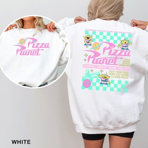 May include: White Pizza Planet sweatshirt with a pink and teal graphic on the front and back. The front features the Pizza Planet logo. The back has the Pizza Planet logo, alien characters, and text that reads "Interstellar Fast-Food Fueling Station."