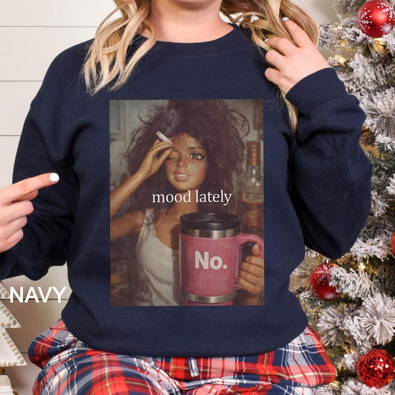 May include: A navy blue crewneck sweatshirt featuring a graphic print of a doll with messy hair holding a cigarette and a pink travel mug with the text No. The text mood lately is printed above the mug. The sweatshirt has long sleeves and a ribbed collar.