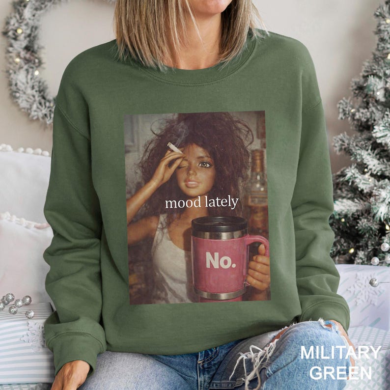 May include: A military green crewneck sweatshirt featuring a graphic print of a doll with messy dark hair holding a cigarette and a pink mug with the text No. The text mood lately is printed above the mug in a white, lowercase serif font.
