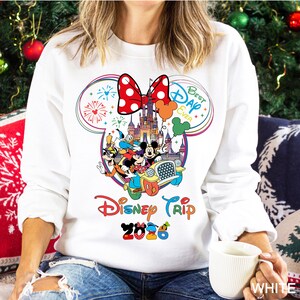 May include: White sweatshirt featuring a colorful Disney graphic. The design includes Mickey Mouse, Donald Duck, and other characters in a car, with text that reads "Disney Trip 2026" and "Best Day Ever". A large red polka dot bow is at the top.