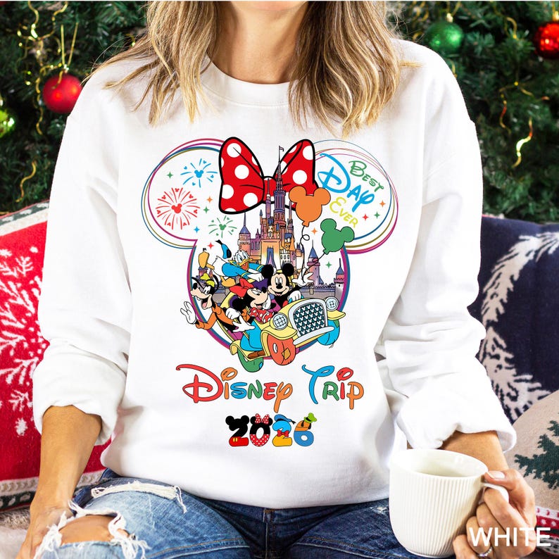 May include: White sweatshirt featuring a colorful Disney graphic. The design includes Mickey Mouse, Donald Duck, and other characters in a car, with text that reads "Disney Trip 2026" and "Best Day Ever". A large red polka dot bow is at the top.