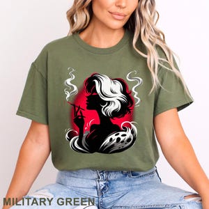 Puede incluir: Camiseta verde militar con un gr&aacute;fico de una mujer fumando, sobre un fondo rojo. La silueta de la mujer es negra, con pelo blanco y negro. El texto "MILITARY GREEN" est&aacute; en la parte inferior de la camiseta.
