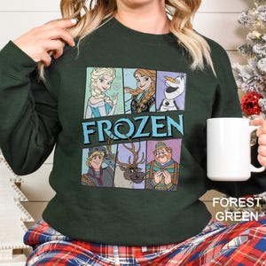 May include: Forest green sweatshirt featuring characters from the movie "Frozen." The design includes Elsa, Anna, Olaf, Kristoff, Sven, and Oaken. The word "FROZEN" is printed in large, blue letters. A white mug is held in the person's hand.