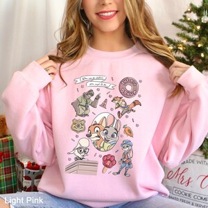 以下が含まれることがあります： 人気アニメ映画のキャラクターと要素のカラフルなグラフィックが特徴のライトピンクのスウェットシャツ。デザインにはドーナツ、警察のバッジ、そして「You can be whatever you wish to be.」というテキストが含まれています。