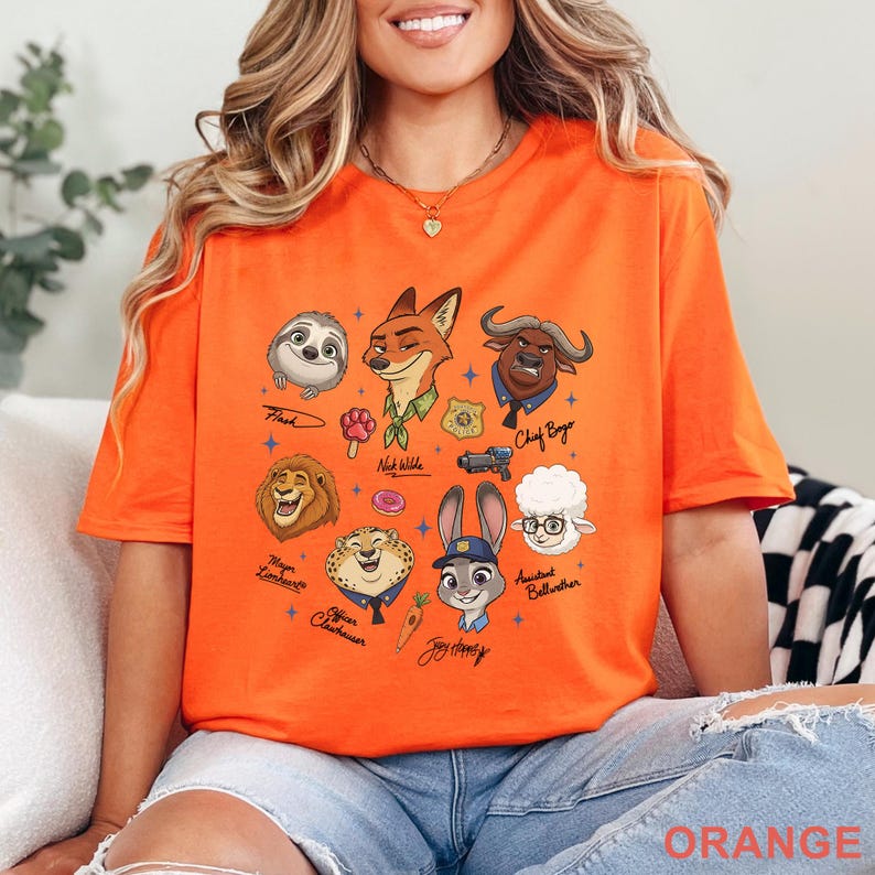 Pu&ograve; includere: T-shirt arancione con personaggi dei cartoni animati di Zootropolis, tra cui Flash, Nick Wilde e Judy Hopps. Il design include nomi e icone dei personaggi. La parola "ORANGE" &egrave; stampata in basso sulla maglietta.