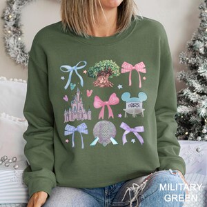 May include: Military green sweatshirt featuring colorful illustrations of bows, a castle, a tree, and other theme park icons. The text "STUDIOS" is visible on a light blue Mickey Mouse head. The bottom right corner of the sweatshirt has the text "MILITARY GREEN".