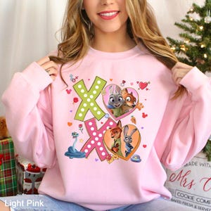 May include: Light pink sweatshirt featuring the characters Judy Hopps and Nick Wilde from Zootopia. The design includes the letters "XO" with heart and dot details, and images of the characters in heart-shaped frames. The text "Light Pink" is at the bottom.