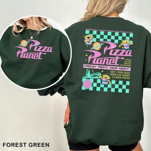 May include: Forest green sweatshirt with the Pizza Planet logo in pink and white. The back features the Pizza Planet logo, aliens, and text that reads "Interstellar Fast-Food Fueling Station" and "Friday Night Kids Night."