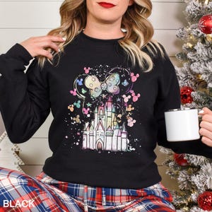 May include: Black sweatshirt featuring a colorful castle design with Mickey Mouse ears and a bow. The design includes pastel colors and a splatter effect. The word "BLACK" is printed at the bottom. A white mug is held in the hand.