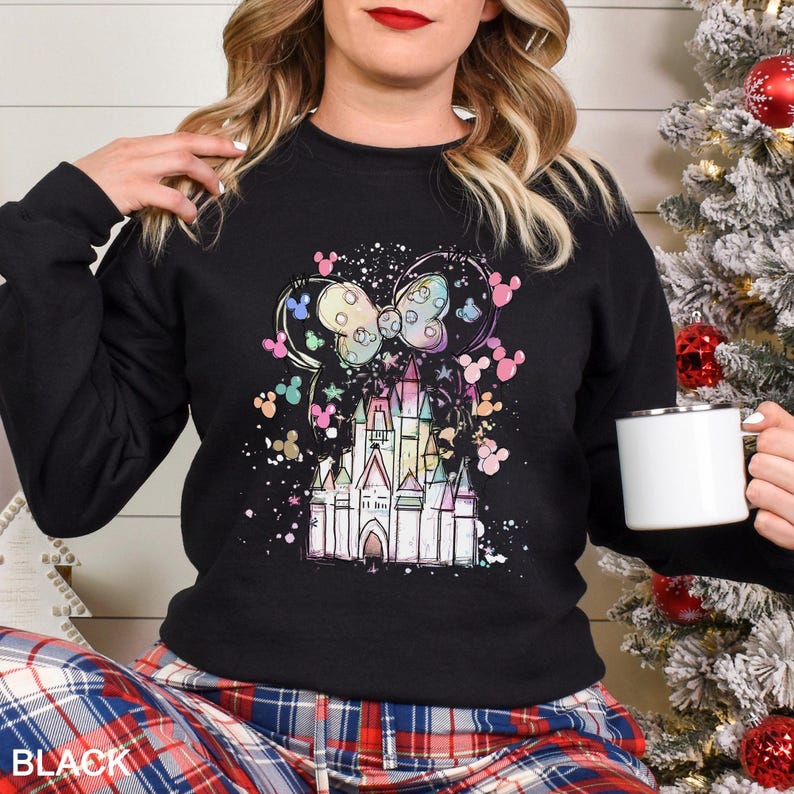 May include: Black sweatshirt featuring a colorful castle design with Mickey Mouse ears and a bow. The design includes pastel colors and a splatter effect. The word "BLACK" is printed at the bottom. A white mug is held in the hand.