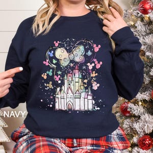 May include: Navy blue sweatshirt featuring a colorful illustration of a castle, Mickey Mouse ears, and a bow. The design includes pastel colors and a splatter effect. The word "NAVY" is printed on the bottom left.