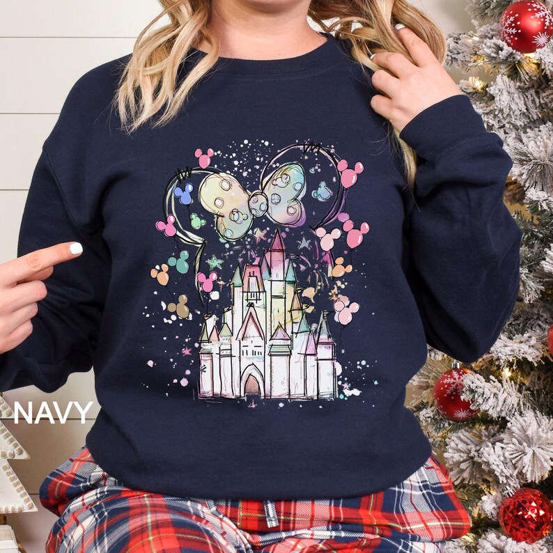 May include: Navy blue sweatshirt featuring a colorful illustration of a castle, Mickey Mouse ears, and a bow. The design includes pastel colors and a splatter effect. The word "NAVY" is printed on the bottom left.