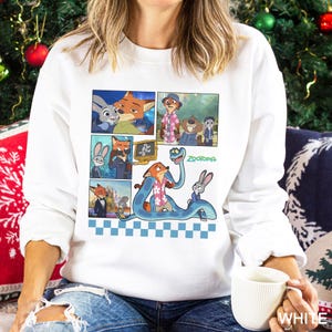 May include: A white long-sleeve sweatshirt featuring a graphic print of various scenes from the movie Zootopia, including characters Nick Wilde and Judy Hopps, with a blue checkered pattern at the bottom and the Zootopia logo in green text.