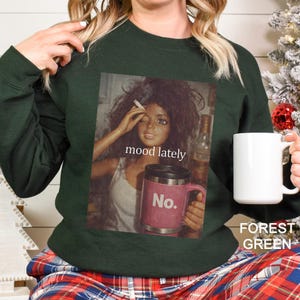 May include: A forest green long-sleeve sweatshirt featuring a graphic print of a doll with messy hair holding a cigarette and a pink travel mug with the text No. The print includes the text mood lately centred above the pink travel mug.