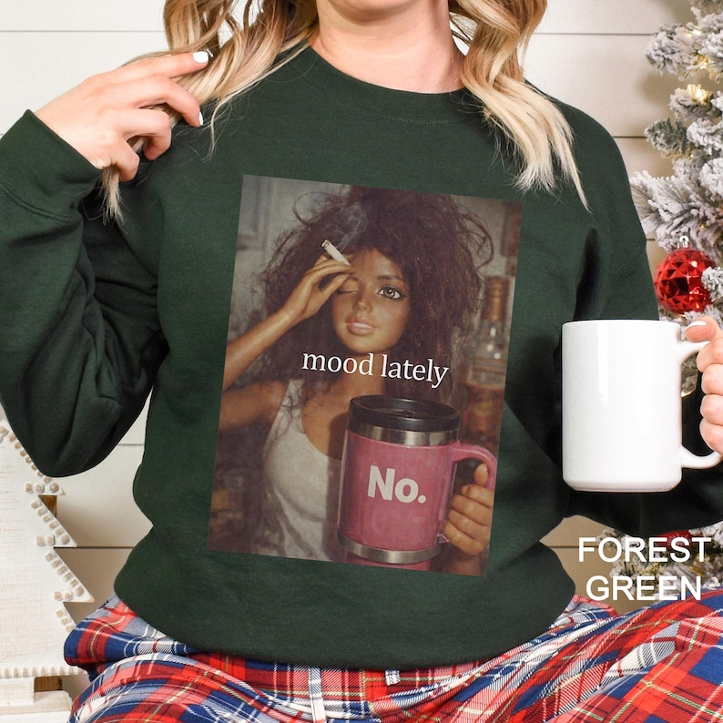 May include: A forest green long-sleeve sweatshirt featuring a graphic print of a doll with messy hair holding a cigarette and a pink travel mug with the text No. The print includes the text mood lately centred above the pink travel mug.