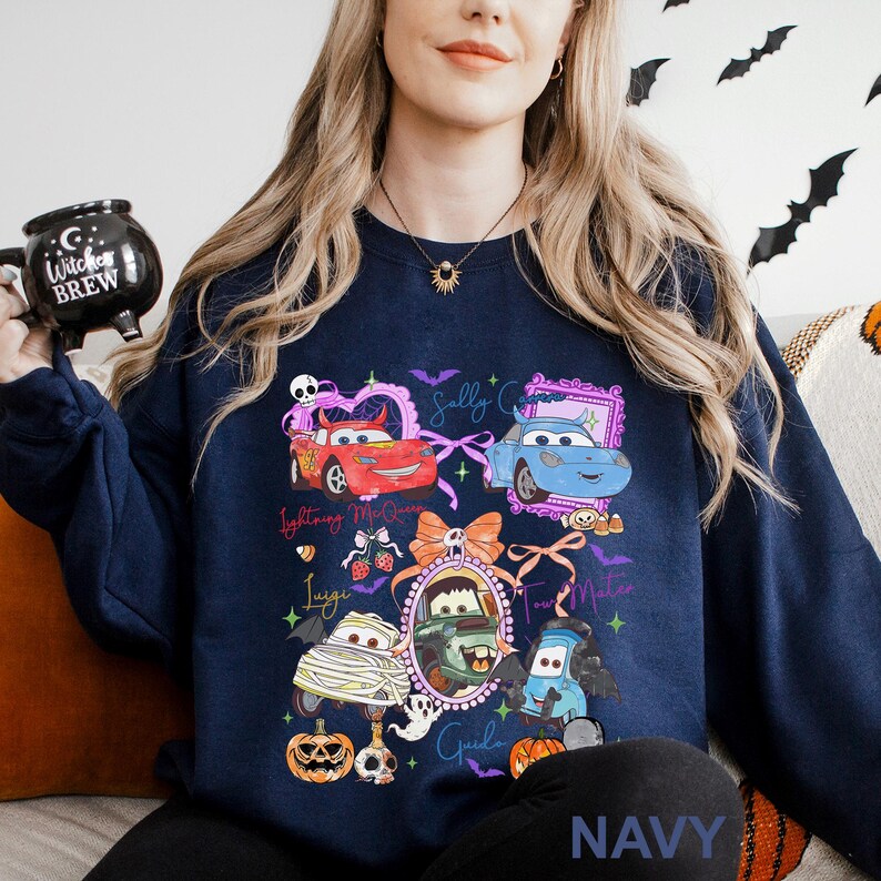 May include: A navy blue long-sleeve sweatshirt featuring a graphic print of various animated car characters in Halloween costumes, including a mummy, a vampire, and a pumpkin, with text labels and decorative elements like bats and bows.