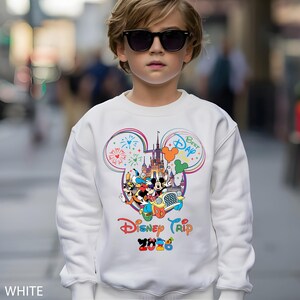 May include: White sweatshirt featuring a colorful Disney Trip 2026 graphic. The design includes Mickey Mouse, Donald Duck, and other characters, with fireworks and a castle. The sweatshirt has a crew neck and long sleeves.