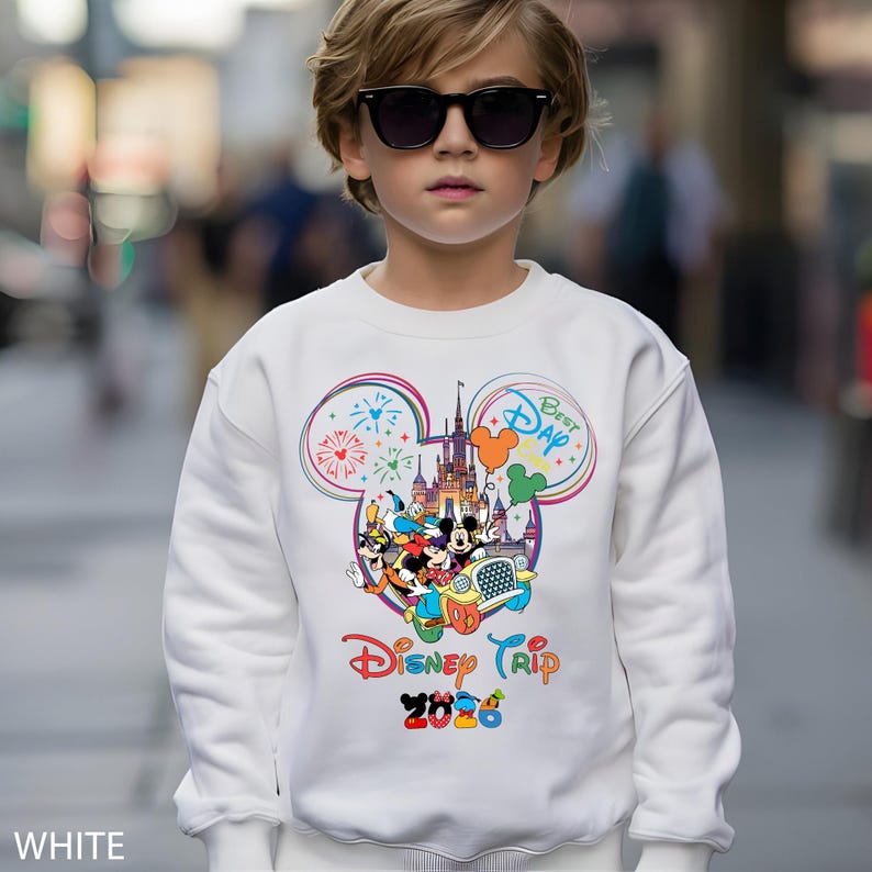 May include: White sweatshirt featuring a colorful Disney Trip 2026 graphic. The design includes Mickey Mouse, Donald Duck, and other characters, with fireworks and a castle. The sweatshirt has a crew neck and long sleeves.