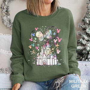 May include: Military green sweatshirt featuring a colorful castle design with a bow and mouse ears. The design includes pastel colors and a sprinkle effect. The sweatshirt is a crew neck style.
