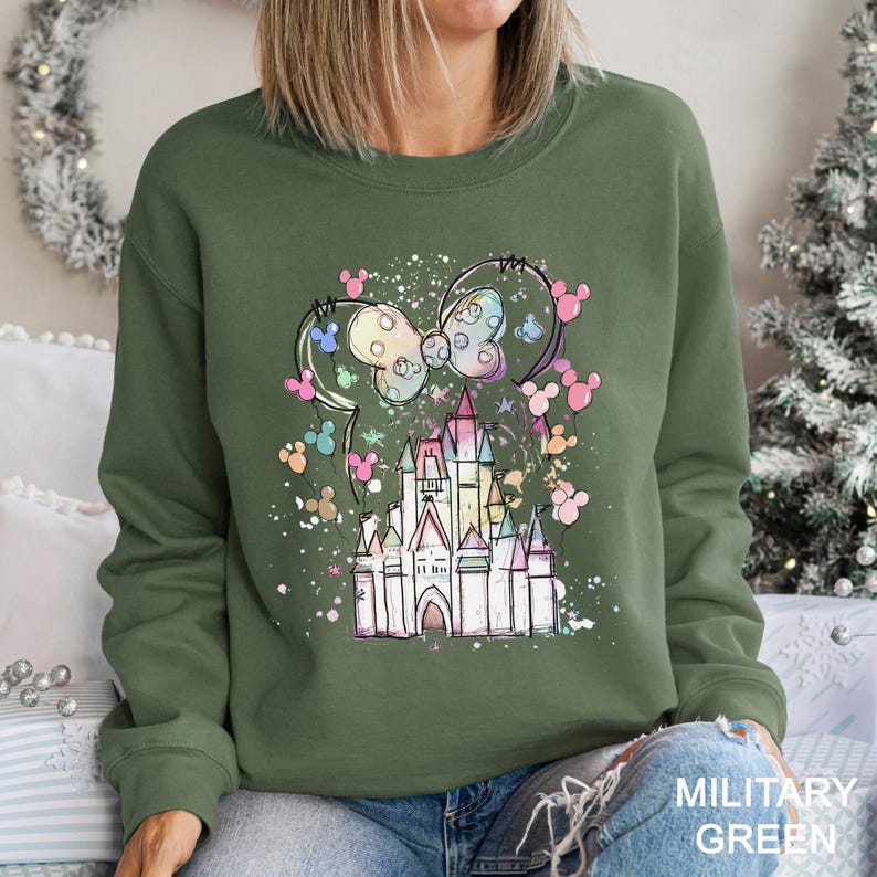 May include: Military green sweatshirt featuring a colorful castle design with a bow and mouse ears. The design includes pastel colors and a sprinkle effect. The sweatshirt is a crew neck style.