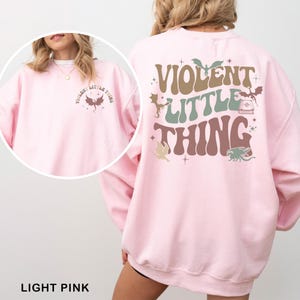 May include: Light pink sweatshirt with the text "VIOLENT LITTLE THING" in brown, green, and tan. The design includes dragon illustrations. The front of the sweatshirt has the same text and dragon graphic.