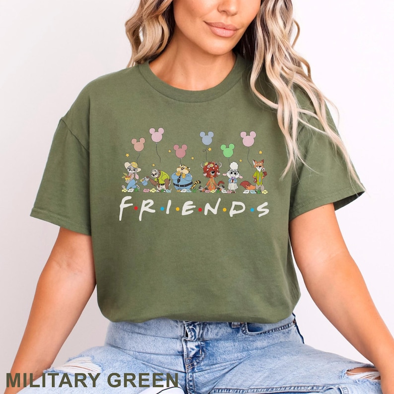 May include: Military green t-shirt featuring a cartoon design of various characters holding Mickey Mouse-shaped balloons. The word "FRIENDS" is printed in colorful letters across the front. The shirt is a comfortable, casual style.