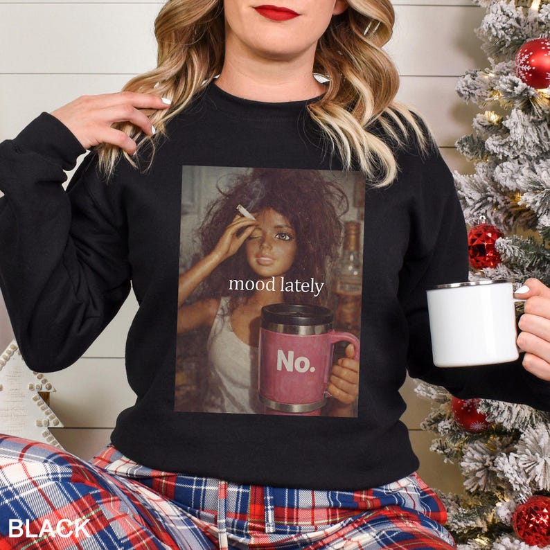 May include: A black crewneck sweatshirt featuring a graphic print of a doll with messy hair holding a cigarette and a pink travel mug with the text No. The text mood lately is printed above the mug. The sweatshirt has long sleeves and a ribbed collar.
