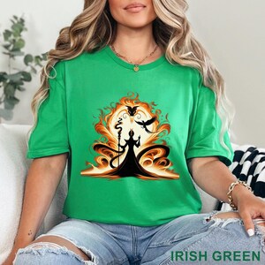 Puede incluir: Camiseta verde irland&eacute;s con un gr&aacute;fico de una figura silueteada con un bast&oacute;n, sobre un sol ardiente con un cuervo. El texto "IRISH GREEN" est&aacute; en la parte inferior. La camiseta es de cuello redondo y manga corta.