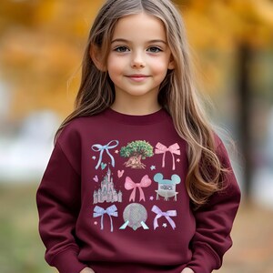 May include: A maroon sweatshirt featuring colorful illustrations of a castle, bows, a tree, and a mouse-eared character. The design is whimsical and playful, perfect for a child's wardrobe. The sweatshirt is made of a soft material.