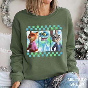 May include: Military green crew neck sweatshirt with a graphic of three cartoon characters: a fox in a pink shirt, a snake, and a rabbit. The graphic is framed with a blue and green checkered pattern. The sweatshirt is a casual style.