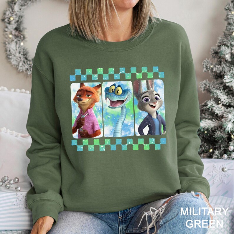 May include: Military green crew neck sweatshirt with a graphic of three cartoon characters: a fox in a pink shirt, a snake, and a rabbit. The graphic is framed with a blue and green checkered pattern. The sweatshirt is a casual style.
