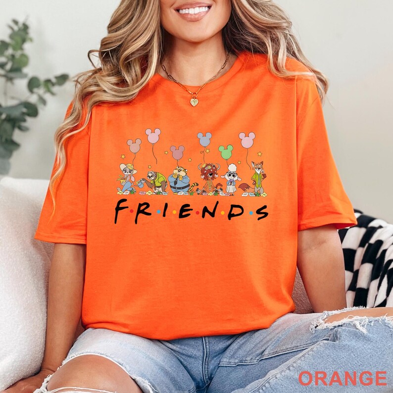 May include: Orange t-shirt featuring the word "FRIENDS" in black, with colorful dots. Above the word are cartoon characters holding Mickey Mouse-shaped balloons. The shirt is a vibrant orange color.