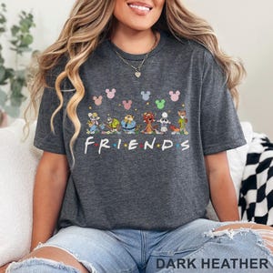 May include: Dark heather gray t-shirt featuring a colorful graphic of cartoon characters and Mickey Mouse heads. The word "FRIENDS" is printed in white letters. The shirt is made of a soft material.