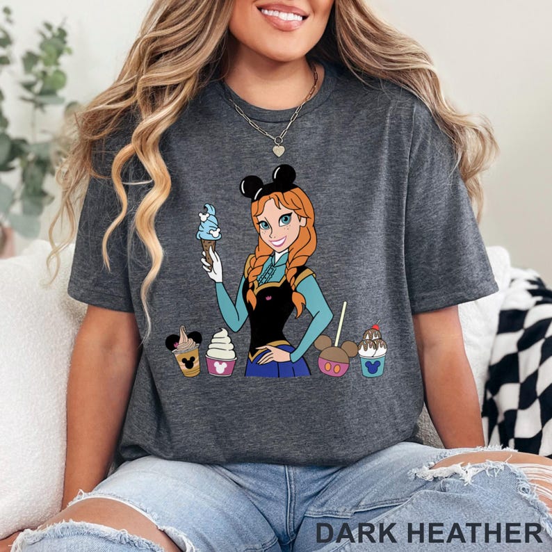 以下が含まれることがあります： ダークヘザーグレーのTシャツ。赤い三つ編みと青いアイスクリームコーンを持ったキャラクターの漫画イラストが描かれています。キャラクターは黒いトップスと青いスカートを着ています。シャツには漫画のカップケーキと「DARK HEATHER」の文字も描かれています。