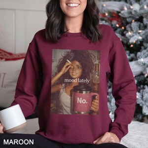 May include: A maroon crewneck sweatshirt featuring a graphic print of a doll with dark hair holding a cigarette and a pink travel mug. The text "mood lately" is printed above the mug, and the word "No." is printed on the mug itself.