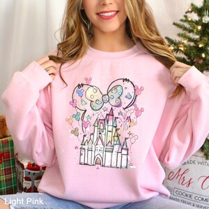 May include: Light pink sweatshirt featuring a whimsical castle design with a large bow and Mickey Mouse ears. The design includes pastel colors and stars. The sweatshirt is a crew neck style.